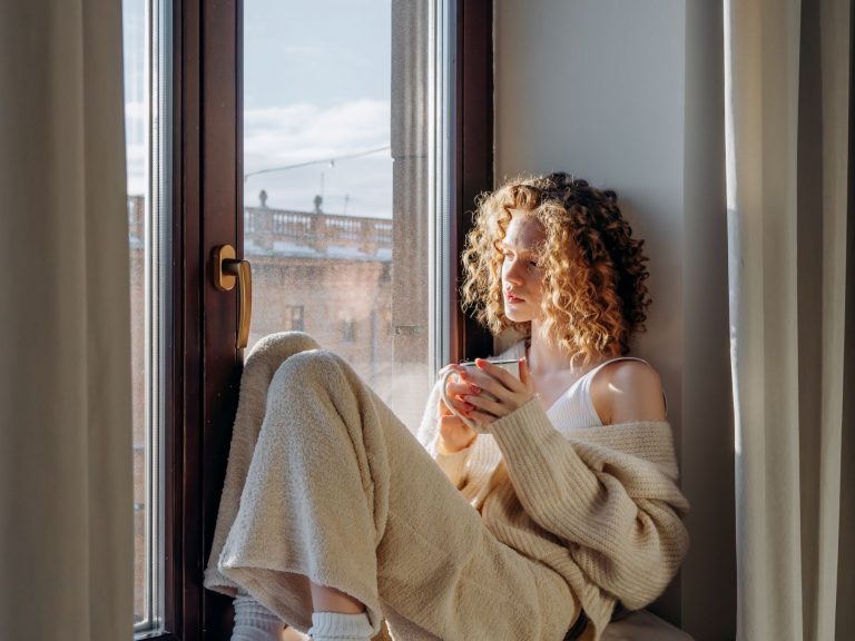 Person sitting by a window holding a cup, reflecting quietly and sensing readiness for a private wellbeing retreat